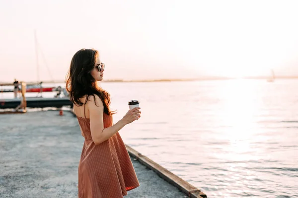 Güzel bir genç kız, siyah bardaklı bir elbiseyle iskelede yürüyor. Deniz tatili, hayat