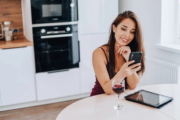 Çekici Asyalı kadın cep telefonuyla şarap içiyor ve evdeki mutfakta tablet kullanıyor.