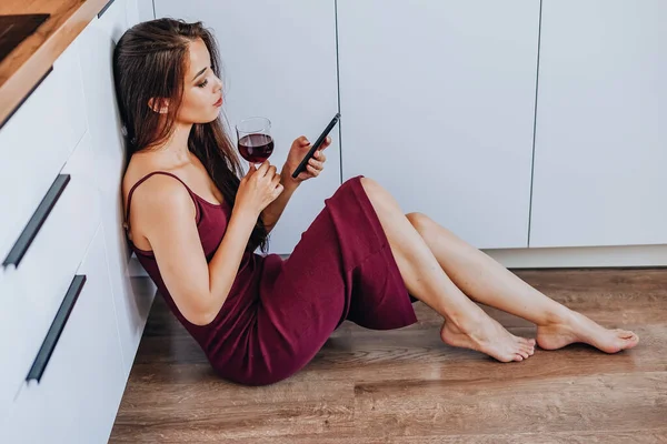 Çekici bir Asyalı kadın şarap içiyor ve evin mutfağında yerde cep telefonu kullanıyor.