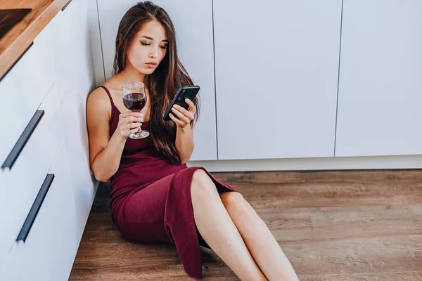 Çekici bir Asyalı kadın şarap içiyor ve evin mutfağında yerde cep telefonu kullanıyor.