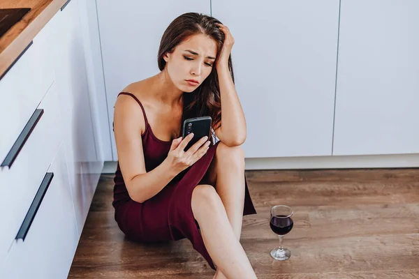 Çekici bir Asyalı kadın şarap içiyor ve evin mutfağında yerde cep telefonu kullanıyor.