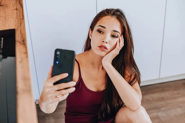 Çekici bir Asyalı kadın şarap içiyor ve evin mutfağında yerde cep telefonu kullanıyor.