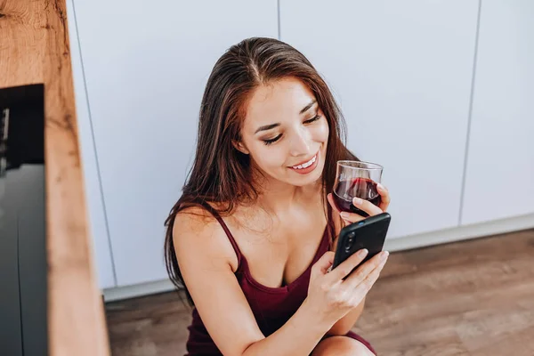 Çekici bir Asyalı kadın şarap içiyor ve evin mutfağında yerde cep telefonu kullanıyor.
