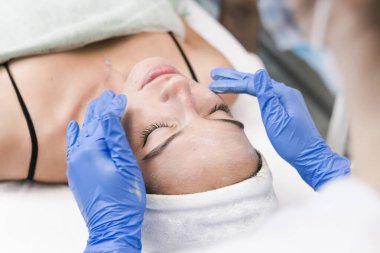 Kozmetik salonunun genç bayan müşterisi yüz maskesi takıyor. Yüzüne soyulan bir maske takma prosedürü. Cilt bakımı ve güzellik salonu veya klinik kavramları.