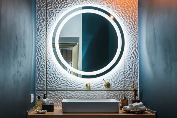 Modern banyo içi beyaz lavabo, altın musluk ve yuvarlak ışıklı ayna..