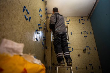 Banyonun tadilatı devam ediyor. İşçi küçük bir merdivenin üzerinde ve banyonun duvarlarına büyük sarı fayanslar yerleştiriyor..