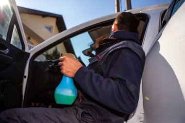 Çalışkan bir işçi beyaz bir arabayı yıkamayı yeni bitiriyor. Şimdi iç kısımları özel bir bez ile siliyor, ve diğer elinde yıkama sıvısı olan bir sprey var..