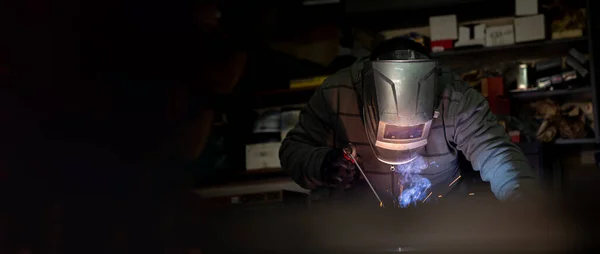 An adult male works in an old workshop. He holds a welding machine in his hand and welds the metal structure. Copy space.