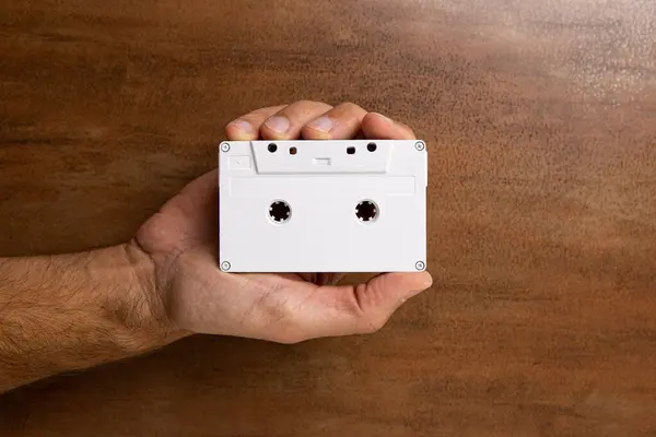 stock image There is a white audio magnetic cassette in the man's hand. Below is a wooden brown background.