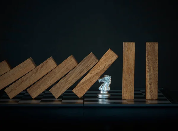 Crisis management, risk management, crisis solving or problem solving concept. Knight chess stopping wooden dominoes from collapsing on chessboard in black scene.