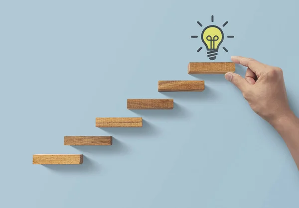 Business growth, business success or career path success concept. Hand is arranging wooden blocks in a shape of staircase on blue background with light bulb icon.