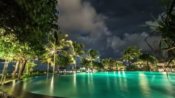 Des nuages flottent au-dessus de la piscine nocturne éclairée sur fond de palmiers et de mer. 4K Timelapse - Bali, Indonésie, juin 2016 .