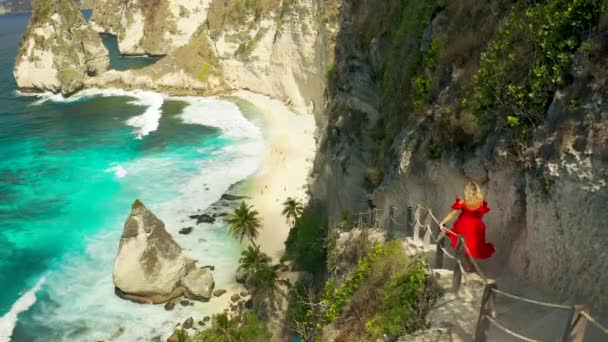 Une jeune femme blonde descend sur une plage tropicale dans les escaliers creusés dans un rocher à la Diamond Beach de l'île de Nusa Penida, Bali, Indonésie. Vue aérienne 4K — Video