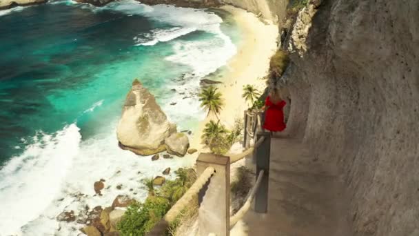 印尼巴厘岛努沙佩尼达岛的钻石海滩上，一名年轻的金发女子走下楼梯，跌落在一块岩石中的热带海滩上。Aerial view 4K — 图库视频影像