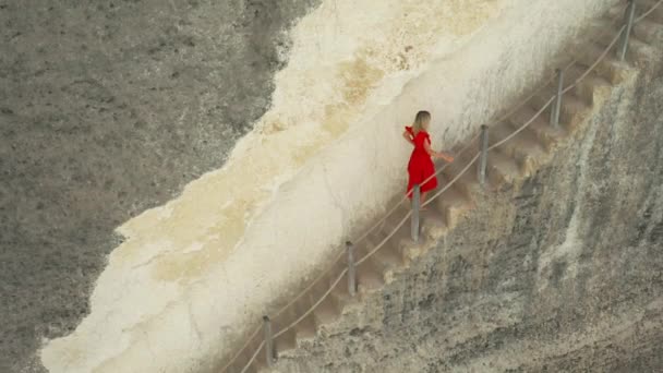 Ung kvinna i röd klänning klättrar en brant trappa nedskuren i en klippa vid Diamond Beach, Nusa Penida Island, Bali, Indonesien. Flygvy 4K — Stockvideo