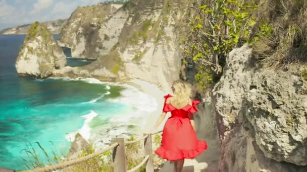 Una joven rubia con un vestido rojo está bajando por las escaleras en la roca que va a la playa del diamante en la isla de Nusa Penida, Bali, Indonesia. — Vídeos de Stock