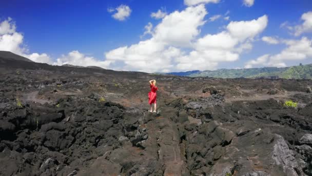 Mujer vestida de rojo en el paisaje negro de los campos de lava en el volcán Batur en Bali, Indonesia. Vista aérea 4K — Vídeo de stock