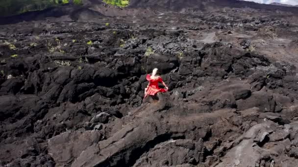 Bali, Endonezya 'daki volkan Batur' da katılaşmış siyah lav tarlasının arka planındaki kırmızı elbiseli genç kadın. Hava görüntüsü 4K — Stok video