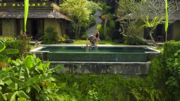 Feliz familia sentada en el borde de la piscina infinita en un paraíso tropical en Ubud, Bali, Indonesia. Anhera Suite Spa, Ubud, Bali, Indonesia - 15 julio 2019. — Vídeos de Stock