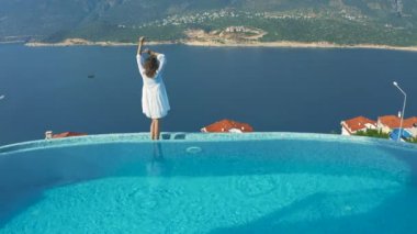 Beyaz elbiseli güzel kadın havuz kenarında duruyor ve Akdeniz 'in yaz manzarasının tadını çıkarıyor. Kas, Hindi. Hava görüntüsü 4K.