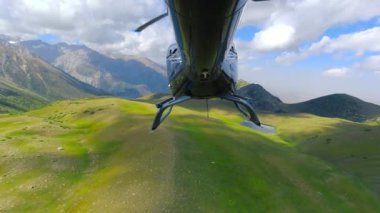 Kuyruk kısmından helikopter görüntüsü kayalık dağlar ve yeşil çayırlar üzerinde uçuyor..
