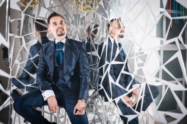 Mavi paltolu yakışıklı adam, aynalarla çevrili bir bar taburesinde oturuyor.