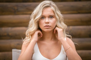 Beyaz elbiseli, sarı dalgalı saçlı güzel kız ahşap duvardaki kameraya bakıyor.