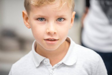 Beyaz tişörtlü, sarı saçlı çocuk kameraya bakıyor.