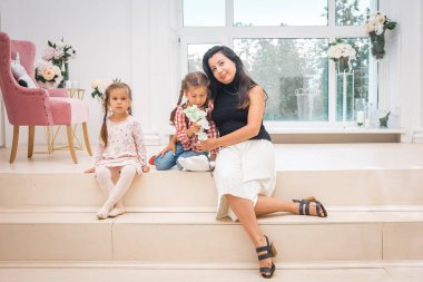 Anne ve iki kızı açık bir iç mekanda büyük bir pencerenin önünde oturuyorlar..