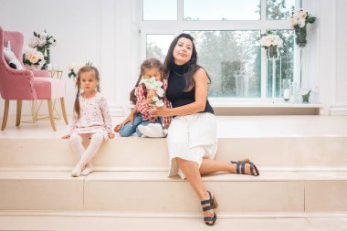 Anne ve iki kızı açık bir iç mekanda büyük bir pencerenin önünde oturuyorlar..