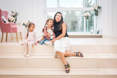 Anne ve iki kızı açık bir iç mekanda büyük bir pencerenin önünde oturuyorlar..