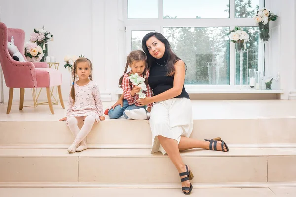 Anne ve iki kızı açık bir iç mekanda büyük bir pencerenin önünde oturuyorlar..