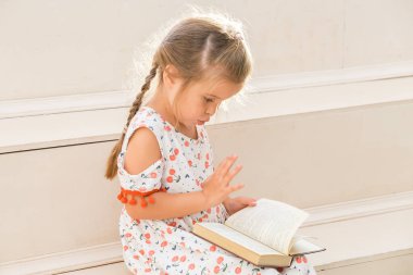 Küçük kız merdivenlerde oturuyor ve büyük bir kitap okuyor..