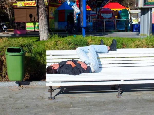 UKRAINE, YALTA - 29 Temmuz 2015: Kotlu ve siyah ceketli evsiz adam sokakta yeşil çöp tenekesinin yanındaki beyaz bankta uyuyor.