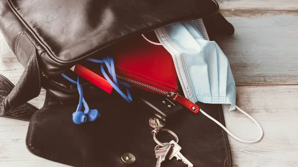 Woman hand bag dropped on wooden floor and open, with accessories and surgical mask. Covid-19 concept.