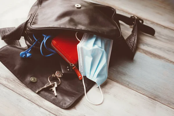 Woman hand bag dropped on wooden floor and open, with accessories and surgical mask. Covid-19 concept.