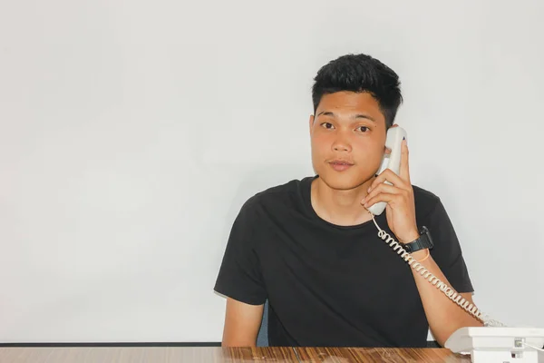 Asyalı adam ahşap masadaki işiyle ilgili konuşmak için akıllı telefon ya da telefon tutuyor, yakışıklı adam beyaz arka planda cep telefonu ya da cep telefonuyla konuşuyor.