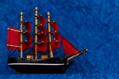 Kırmızı yelkenli siyah tahta bir oyuncak gemi, mavi kağıda vurularak denizi ve gökyüzünü yaratıyor.