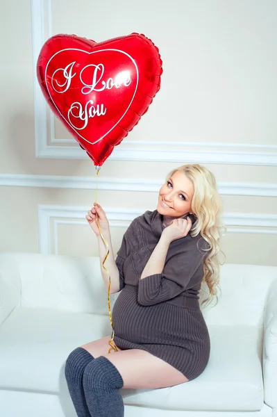 Mladá krásná těhotná žena sedí na gauči a llooking na balónek v podobě srdce. — Stock fotografie