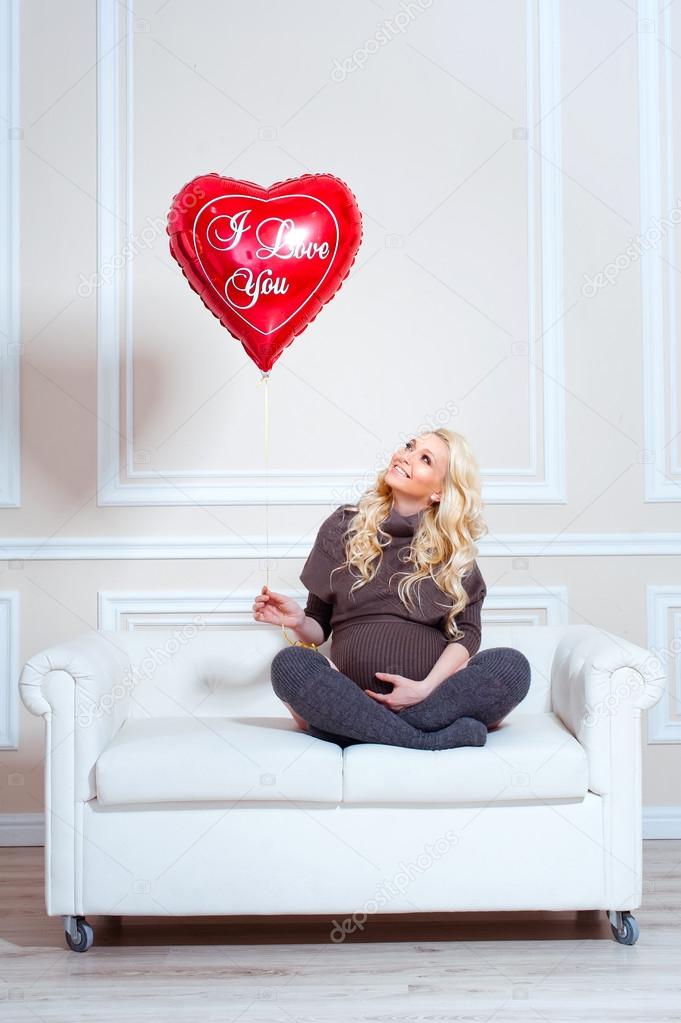 Young beautiful pregnant woman sitting on sofa and llooking at the balloon in the form of heart.