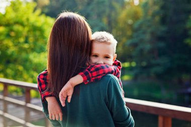 Kadın arkası dönük durur ve kucaklayan çocuğun oğlunu tutar. Bir anne bir yaz günü parkta bir çocuğa sarılır. Mutlu otantik aile çocukluk hayatı.