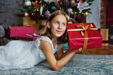 A girl in a white dress is lying at the Christmas tree with a gift in her hands. He looks away dreamily. clipart