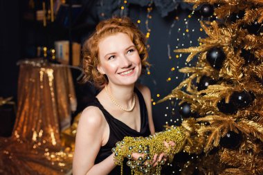 Genç bir kadın, elinde altın süslemelerle bir Noel ağacının arka planında boncuk dizisi tutuyor. Şık kadınlar, şık gece kıyafetleri, aksesuarlar, makyaj. Mutlu yıllar..