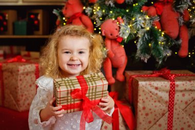 Beyaz elbiseli küçük sarışın kız Noel ağacının arka planında bir hediye ve çelenk ışıkları var..