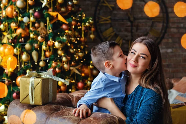 The son kisses his mother on the cheek against the background of a Christmas tree and lights of garlands. Merry Christmas.