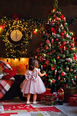 Zarif bir elbisesi olan küçük bir kız Noel ağacını Noel arifesinde evde süslüyor. Kış dekoru olan bir odada bir çocuk Noel ağacına balon asıyor..