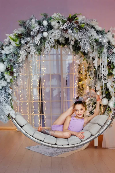 A girl in a gymnastic costume lies on a round swing in a gymnastic pose with christmas decorations in the interior decorated for the new year. The concept of Christmas holidays.