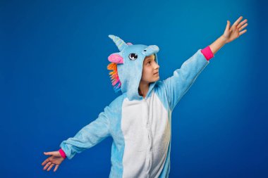 Çekici kız tek boynuzlu at kostümü içinde şakacı poz veriyor. Kigurumi 'deki duygusal bir kadının mavi arka planda eğlenirken çekilmiş bir Stüdyo fotoğrafı. Neşeli bir şekilde ellerini yana açıyor..