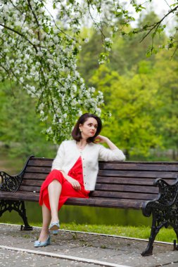 Bahar parkında bankta oturan güzel genç bayanın portresi. Arka planda çiçekler olan çalılar.