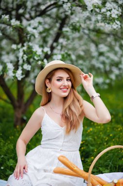 Şapkalı ve elbiseli pozitif bir bayan çiçek açan bir elma ağacının arka planına gülüyor. Bahçede gülümseyen iyimser bir kadın. Bahar geldi.